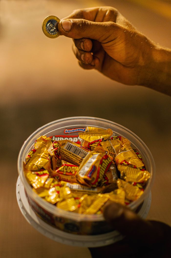 Close-up of a hand with a coin above a box of yellow-wrapped candies at night.